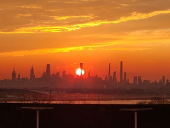 Fiery orange sunset over the New York City skyline, sun dipping between silhouetted skyscrapers with Hudson River reflections and a rooftop foreground.