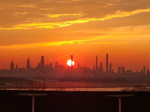 Fiery orange sunset over the New York City skyline, sun dipping between silhouetted skyscrapers with Hudson River reflections and a rooftop foreground.