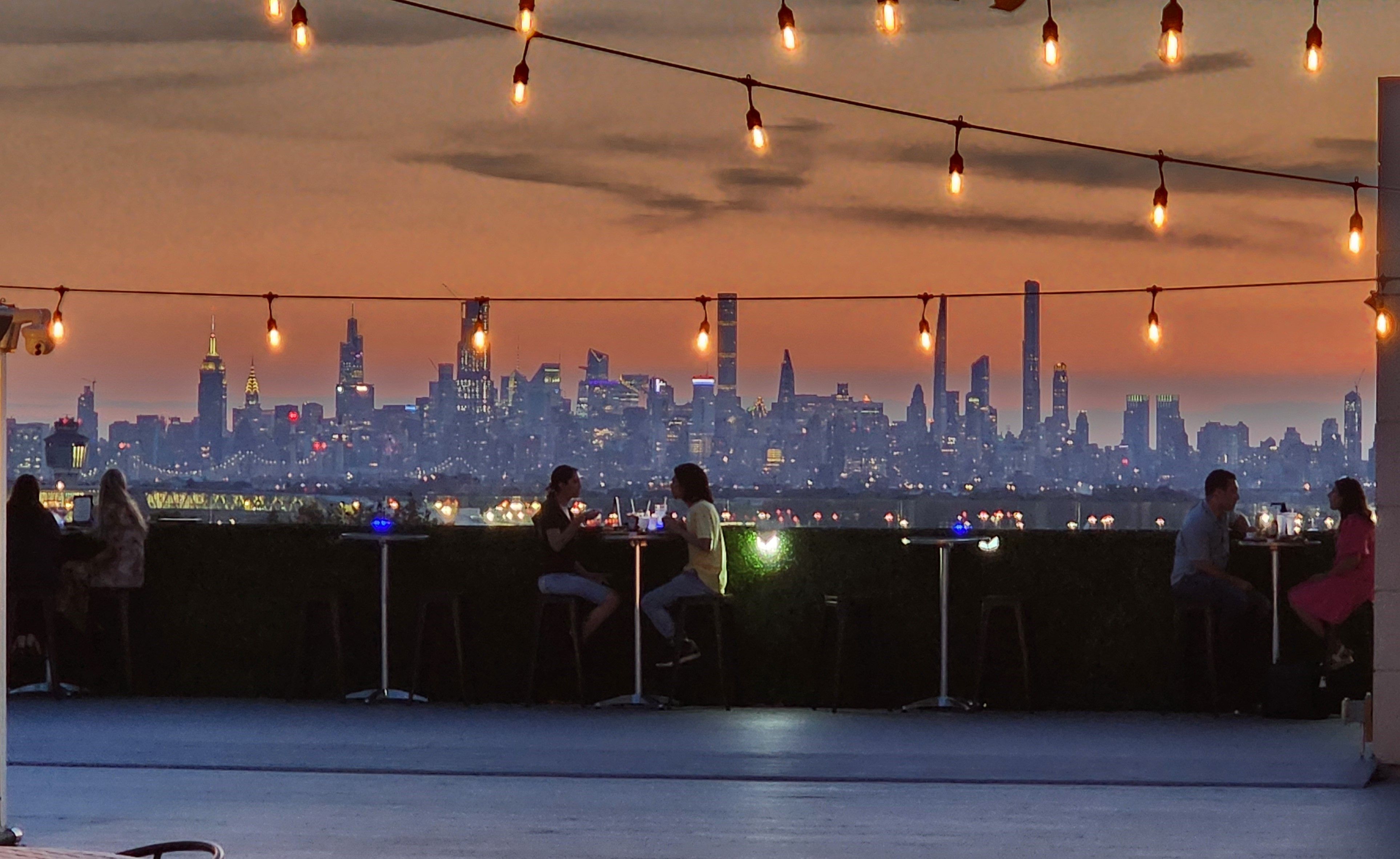 Rooftop terrace at sunset with twinkling string lights and diners at high tables, silhouetted against the Manhattan skyline and glowing city lights.