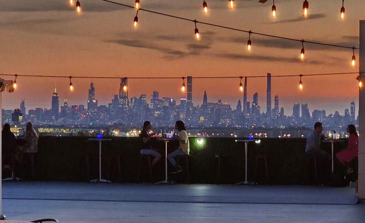 Rooftop terrace at sunset with twinkling string lights and diners at high tables, silhouetted against the Manhattan skyline and glowing city lights.