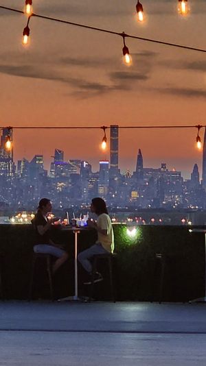 Rooftop terrace at sunset with twinkling string lights and diners at high tables, silhouetted against the Manhattan skyline and glowing city lights.