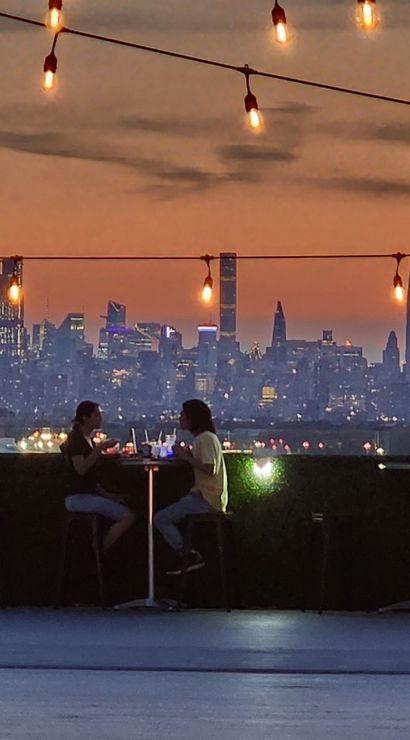 Rooftop terrace at sunset with twinkling string lights and diners at high tables, silhouetted against the Manhattan skyline and glowing city lights.