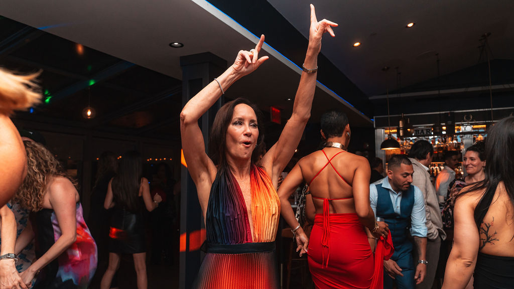 Vibrant indoor dance floor at a lively bar — a woman in a rainbow halter dress raises her arms while others in red and stylish outfits dance around her.