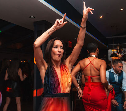 Vibrant indoor dance floor at a lively bar — a woman in a rainbow halter dress raises her arms while others in red and stylish outfits dance around her.