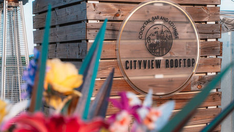 Wooden rooftop cocktail bar sign with city skyline logo on a wood-paneled wall, blurred colorful flowers and spiky foliage in the foreground and a patio heater at left — urban rooftop lounge vibe.