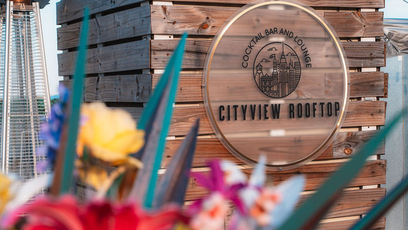 Wooden rooftop cocktail bar sign with city skyline logo on a wood-paneled wall, blurred colorful flowers and spiky foliage in the foreground and a patio heater at left — urban rooftop lounge vibe.