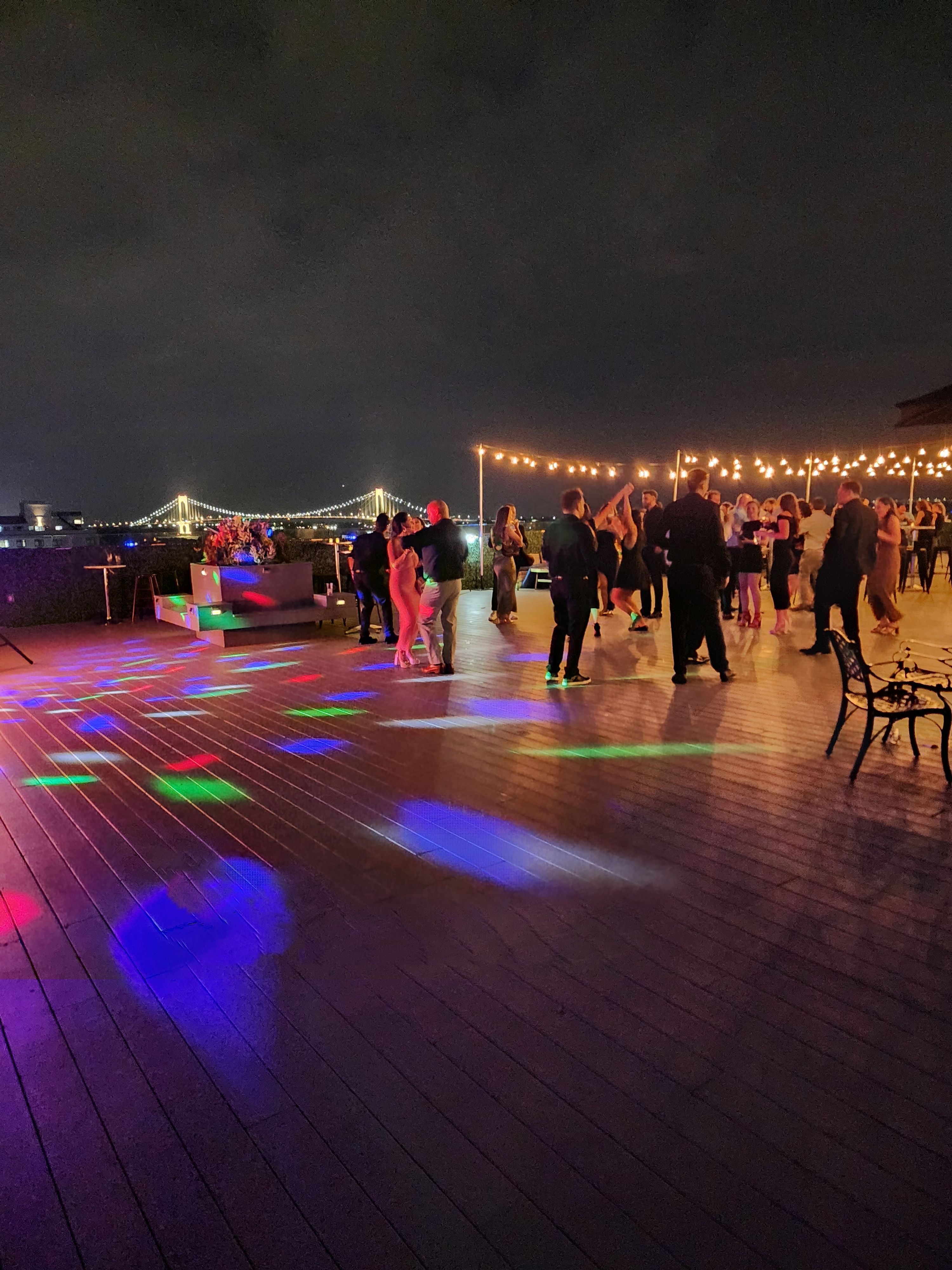 Rooftop waterfront party at night with people dancing under string lights, colorful disco spots, and an illuminated suspension bridge in the skyline.