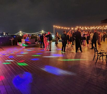 Rooftop waterfront party at night with people dancing under string lights, colorful disco spots, and an illuminated suspension bridge in the skyline.