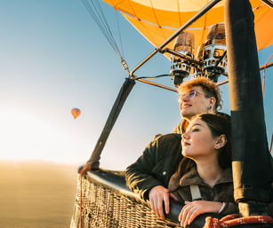 Two people enjoying a sunrise hot air balloon ride over open countryside, gazing from the wicker basket beneath a bright yellow envelope.