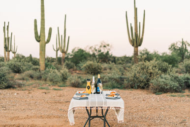 Desert picnic for two in the Sonoran Desert at dusk — small table with white cloth, champagne and flutes, orange juice carafe and pastries, framed by tall saguaro cacti and scrub brush.