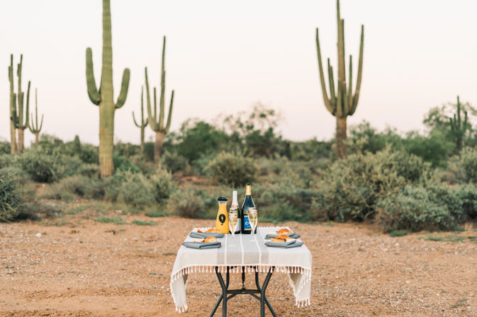 Desert picnic for two in the Sonoran Desert at dusk — small table with white cloth, champagne and flutes, orange juice carafe and pastries, framed by tall saguaro cacti and scrub brush.