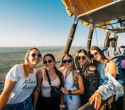 Five smiling women in sunglasses pose together in a hot air balloon basket above wide open plains with distant mountains under a clear blue sky — sunny aerial balloon ride.