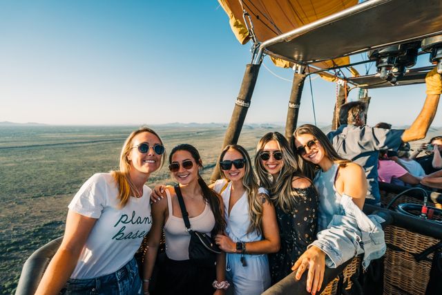 Five smiling women in sunglasses pose together in a hot air balloon basket above wide open plains with distant mountains under a clear blue sky — sunny aerial balloon ride.