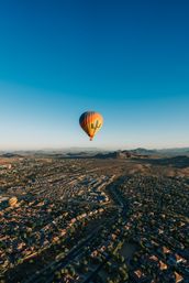 Aerial view of a colorful hot air balloon with a cactus design floating over a desert suburb and rolling foothills at sunrise under a clear blue sky.