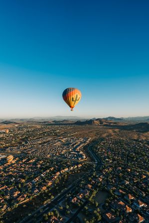 Aerial view of a colorful hot air balloon with a cactus design floating over a desert suburb and rolling foothills at sunrise under a clear blue sky.