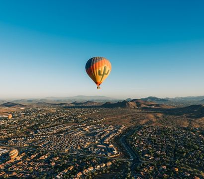 Aerial view of a colorful hot air balloon with a cactus design floating over a desert suburb and rolling foothills at sunrise under a clear blue sky.