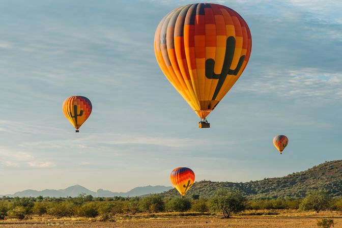 Vibrant orange-and-red hot air balloons with cactus silhouettes drifting over a sunlit Southwestern desert landscape of scrub and distant mountains.