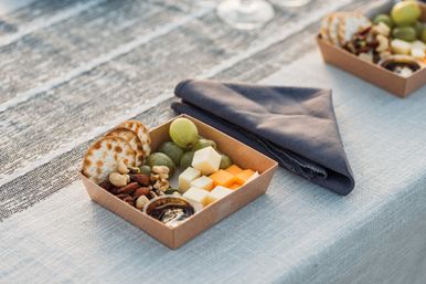 Outdoor picnic charcuterie box with crackers, green grapes, mixed nuts, white and orange cheese cubes and dipping sauce on a striped tablecloth next to a folded navy napkin