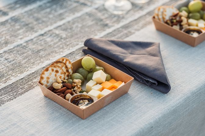 Outdoor picnic charcuterie box with crackers, green grapes, mixed nuts, white and orange cheese cubes and dipping sauce on a striped tablecloth next to a folded navy napkin