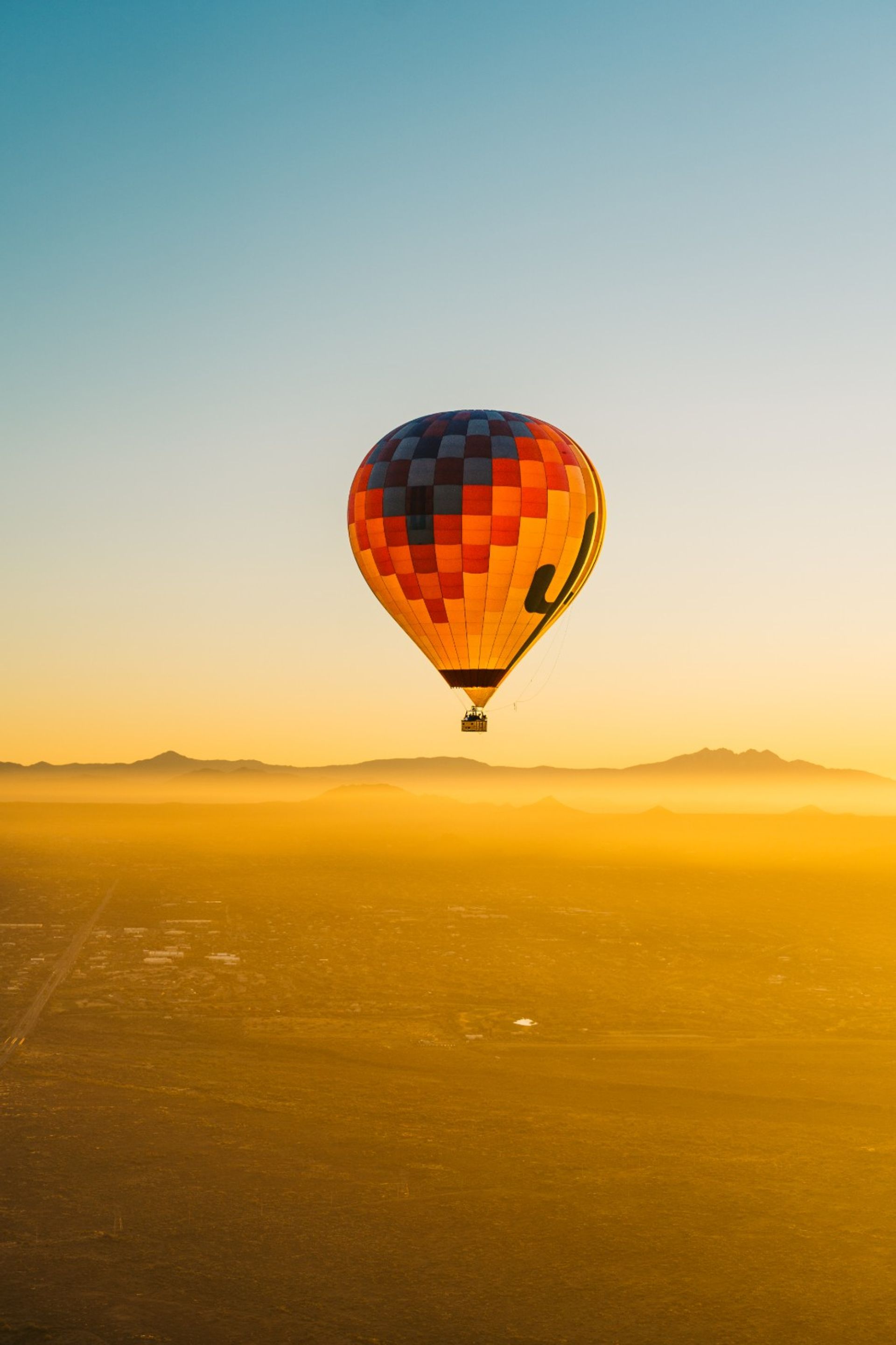 Colorful checkered hot air balloon drifting over a golden desert valley at sunrise with layered mountains on the horizon
