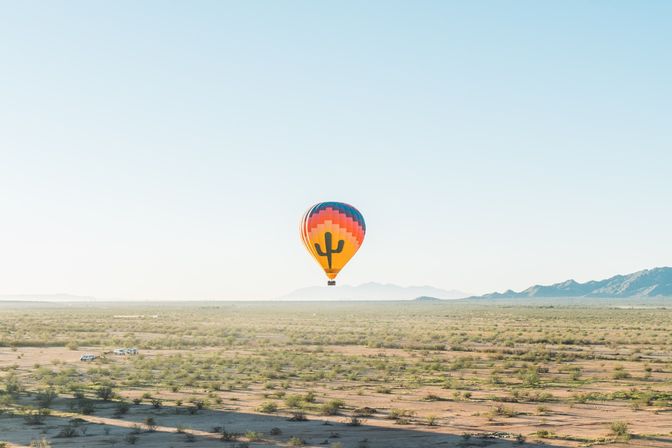 Colorful hot air balloon with a cactus graphic floating over a sunlit desert plain dotted with shrubs and a distant mountain range under a clear blue sky.