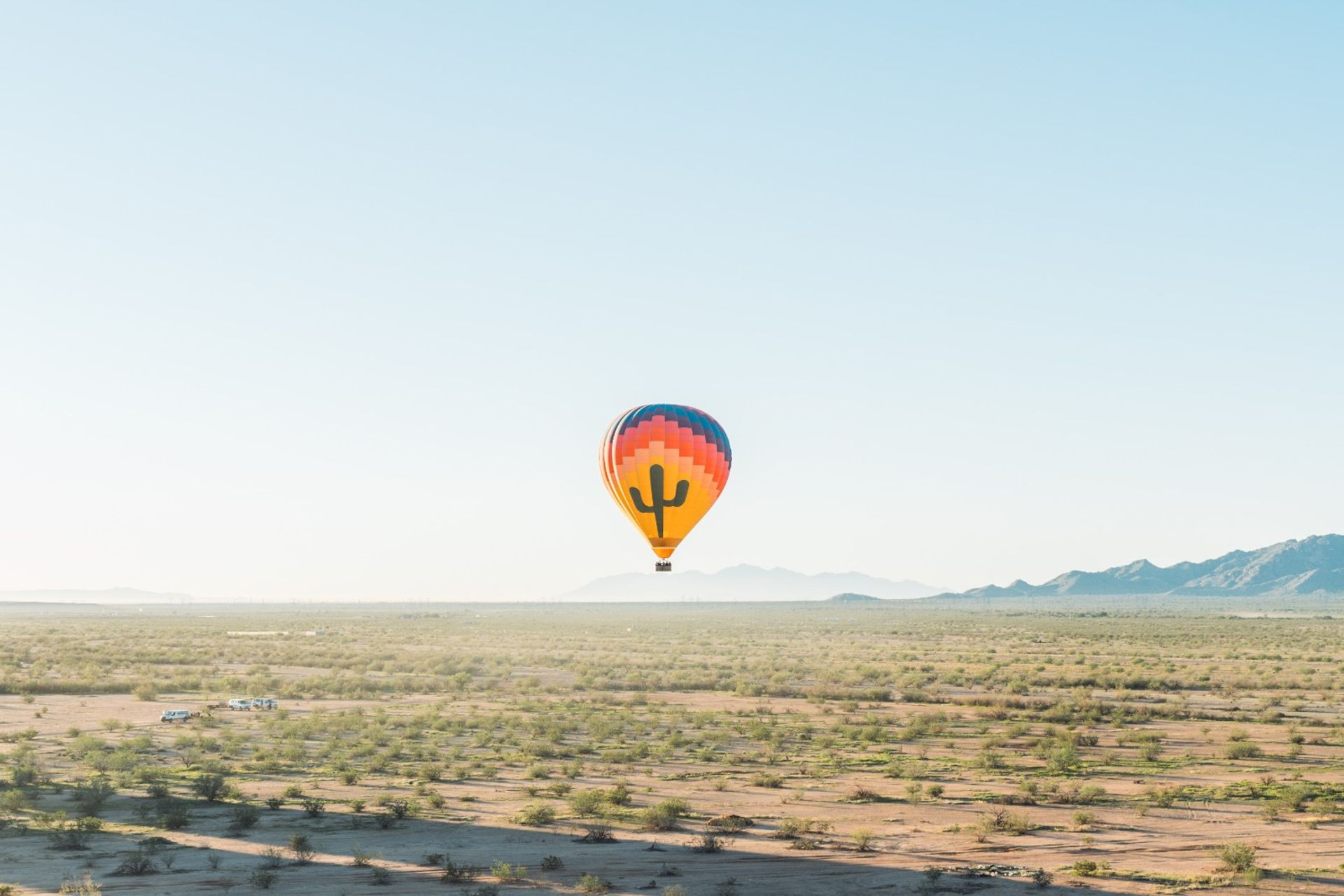 Colorful hot air balloon with a cactus graphic floating over a sunlit desert plain dotted with shrubs and a distant mountain range under a clear blue sky.