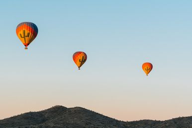 Three colorful hot air balloons with cactus designs drifting over rocky desert hills at sunrise under a clear blue sky