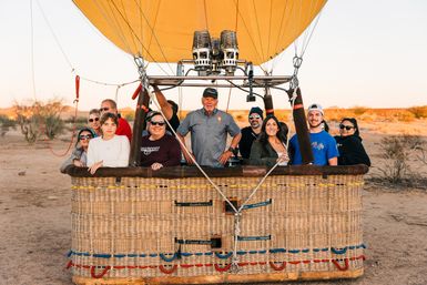 Group of smiling passengers and a pilot posing in a wicker hot air balloon basket under a yellow envelope at sunrise over a desert landscape — scenic hot air balloon ride adventure.