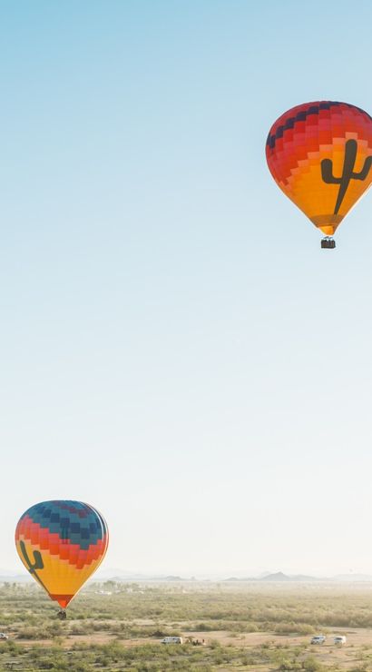 Two colorful hot air balloons with cactus silhouettes floating over a sunlit Southwestern desert plain with distant mountains under a clear blue sky