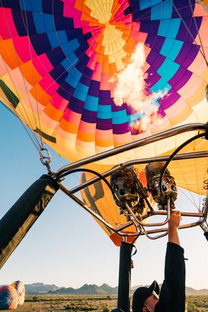 Vibrant hot air balloon canopy and burner flame seen from the basket during a sunrise ride over a desert plain with distant mountain range.