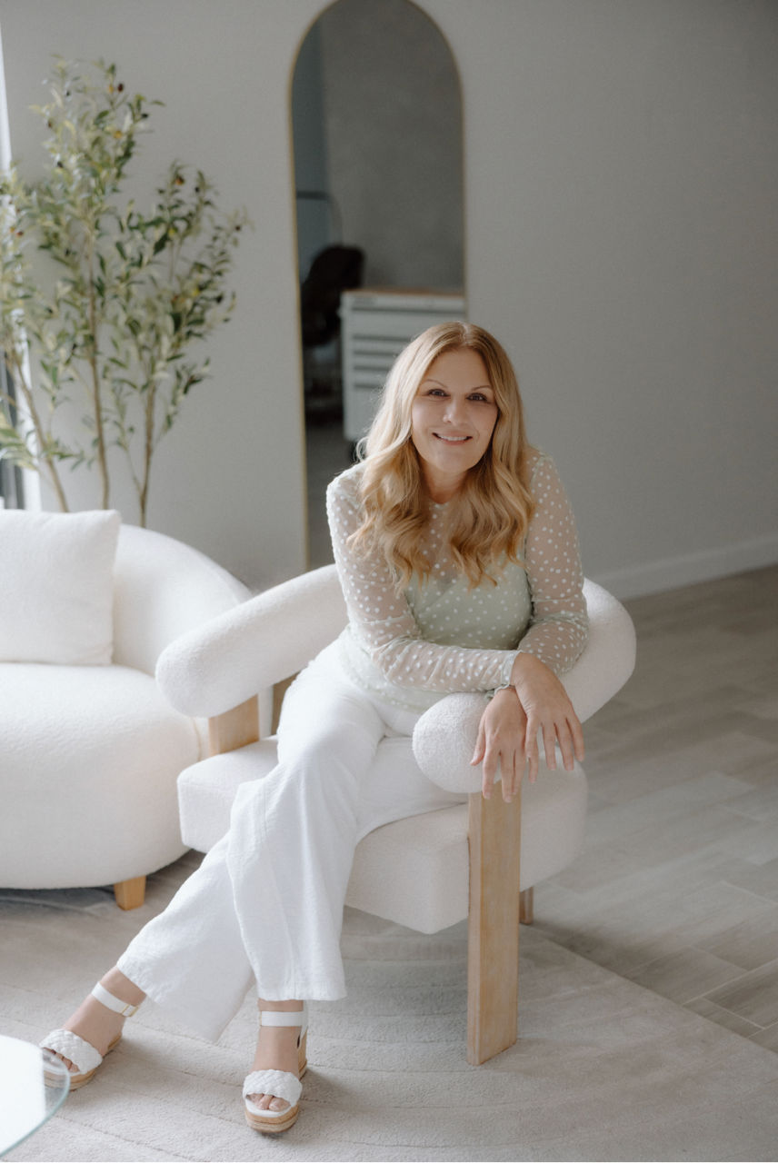 Smiling woman with blonde hair seated on a boucle armchair in a bright, minimalist neutral living room, wearing a sheer polka-dot top and white pants with an arched mirror, potted plant, and light wood floors in the background.