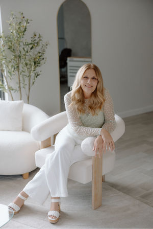 Smiling woman with blonde hair seated on a boucle armchair in a bright, minimalist neutral living room, wearing a sheer polka-dot top and white pants with an arched mirror, potted plant, and light wood floors in the background.