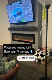 Cozy living room with electric fireplace and TV showing a "Welcome to Watkins Glen" sign; a person reclines on a couch with an IV drip bag nearby and on-screen text promoting IV therapy.