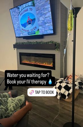 Cozy living room with electric fireplace and TV showing a "Welcome to Watkins Glen" sign; a person reclines on a couch with an IV drip bag nearby and on-screen text promoting IV therapy.