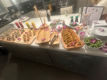 Fun sushi catering spread on a modern kitchen island: wooden boat platters and boards piled with assorted sushi rolls and bites, edamame bowls, soy bottles, condiments, chopsticks and purple orchid garnishes.