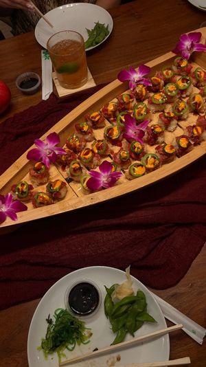 Wooden sushi boat of bite-sized spicy tuna crispy rice topped with jalapeño slices, creamy orange sauce and purple orchid garnishes on a wooden table with edamame, seaweed salad, soy sauce and a cocktail.