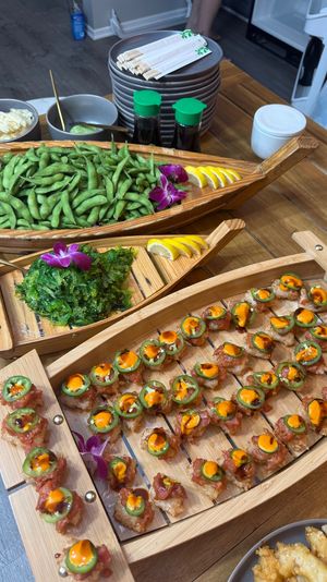 Wooden sushi boat platters on a table with bite-size spicy tuna rice crisps topped with jalapeño and orange sauce, a boat of fresh edamame, seaweed salad, lemon wedges and soy sauce bottles