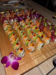 Close-up sushi platter on a wooden board with assorted rolls — shrimp and avocado rolls topped with spicy mayo dots, slices of salmon and tuna, and a purple orchid garnish.