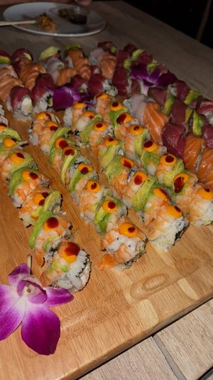 Close-up sushi platter on a wooden board with assorted rolls — shrimp and avocado rolls topped with spicy mayo dots, slices of salmon and tuna, and a purple orchid garnish.