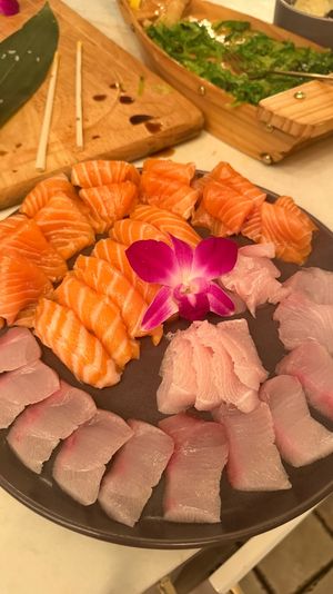 Mouthwatering sashimi platter of orange salmon nigiri and white fish slices arranged on a dark plate with a purple orchid garnish, chopsticks and a wooden board with seaweed salad in the background.