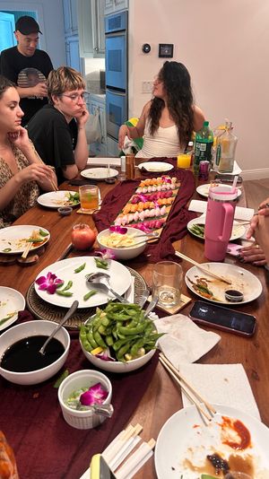 Casual home sushi feast with friends around a wooden dining table — colorful nigiri lined on a cloth runner, edamame, soy sauce bowls, drinks and orchid garnishes in a cozy kitchen setting.