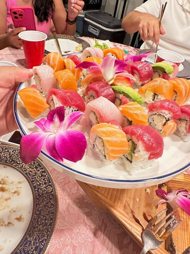 Colorful rainbow sushi platter with salmon, tuna and avocado rolls, purple orchid garnishes and chopsticks on a festive dinner party table with red cups