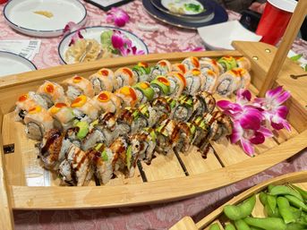 Assorted sushi rolls (avocado, shrimp, eel sauce) arranged on a wooden boat platter with purple orchid garnishes, edamame and place settings on a pink tablecloth — colorful party-style sushi spread.