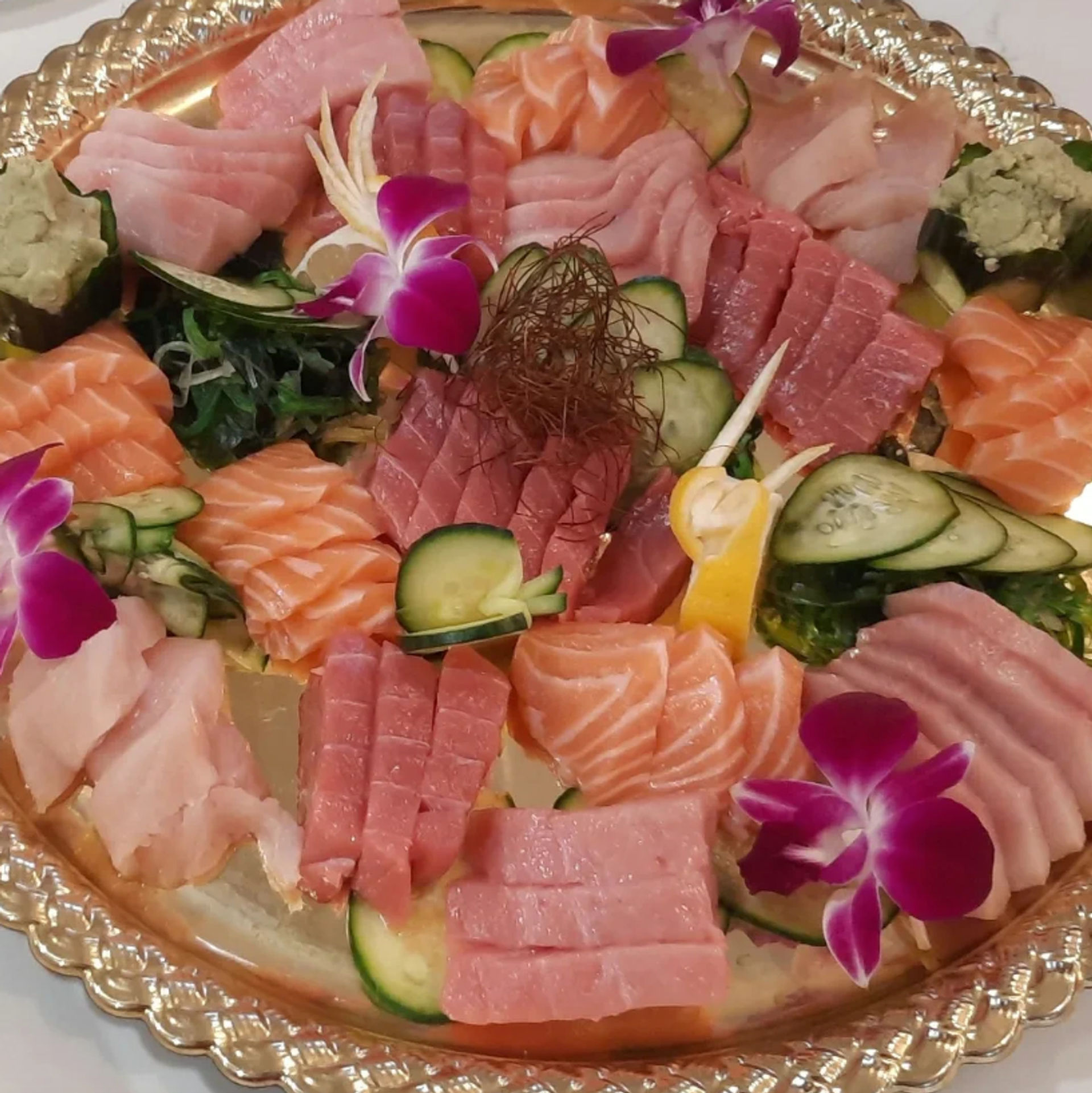 Vibrant Japanese sashimi platter on a gold tray with sliced salmon, tuna and whitefish, cucumber, seaweed salad, wasabi, lemon and purple orchid garnishes