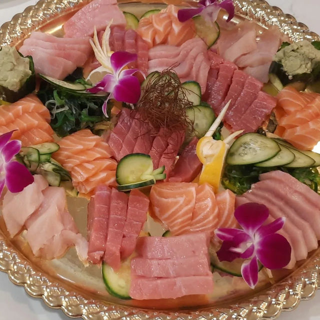 Vibrant Japanese sashimi platter on a gold tray with sliced salmon, tuna and whitefish, cucumber, seaweed salad, wasabi, lemon and purple orchid garnishes