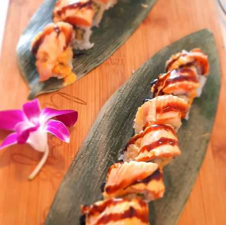 Seared salmon sushi roll drizzled with dark eel sauce, served on green bamboo leaves atop a wooden board with a purple orchid garnish.
