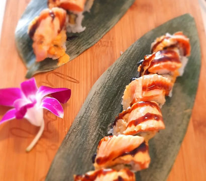 Seared salmon sushi roll drizzled with dark eel sauce, served on green bamboo leaves atop a wooden board with a purple orchid garnish.