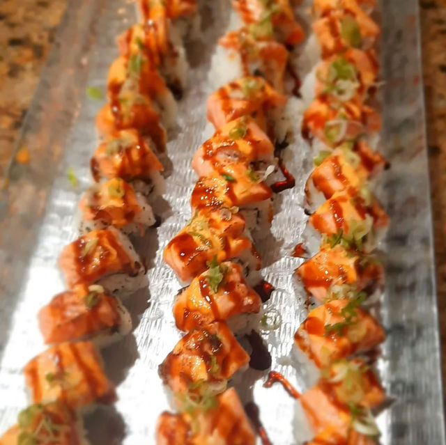 Close-up of three rows of zesty spicy salmon sushi rolls on a metal serving tray, topped with creamy spicy mayo, eel sauce drizzle and chopped green onions.