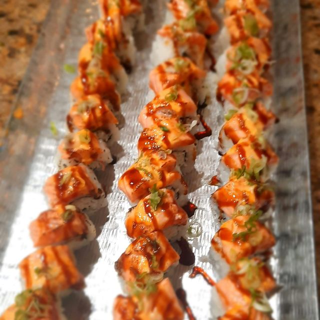 Vibrant close-up of three rows of salmon-topped sushi rolls on a metal platter, drizzled with spicy mayo and eel sauce and sprinkled with green onions.