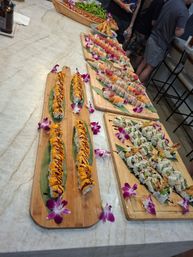 Vibrant sushi roll spread on wooden boards — salmon, tuna and shrimp tempura rolls drizzled with spicy mayo and eel sauce, garnished with purple orchid flowers on a marble countertop.